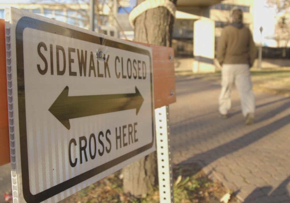 A man walking on a sidewalk near a "Sidewalk Closed, Cross Here" sign.