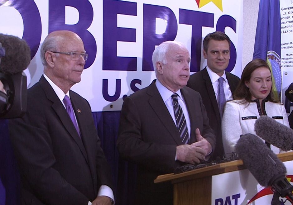 Photo of Pat Roberts, John McCain, Kevin Yoder and Yoder's wife standing behind a podium surrounded by cameras.