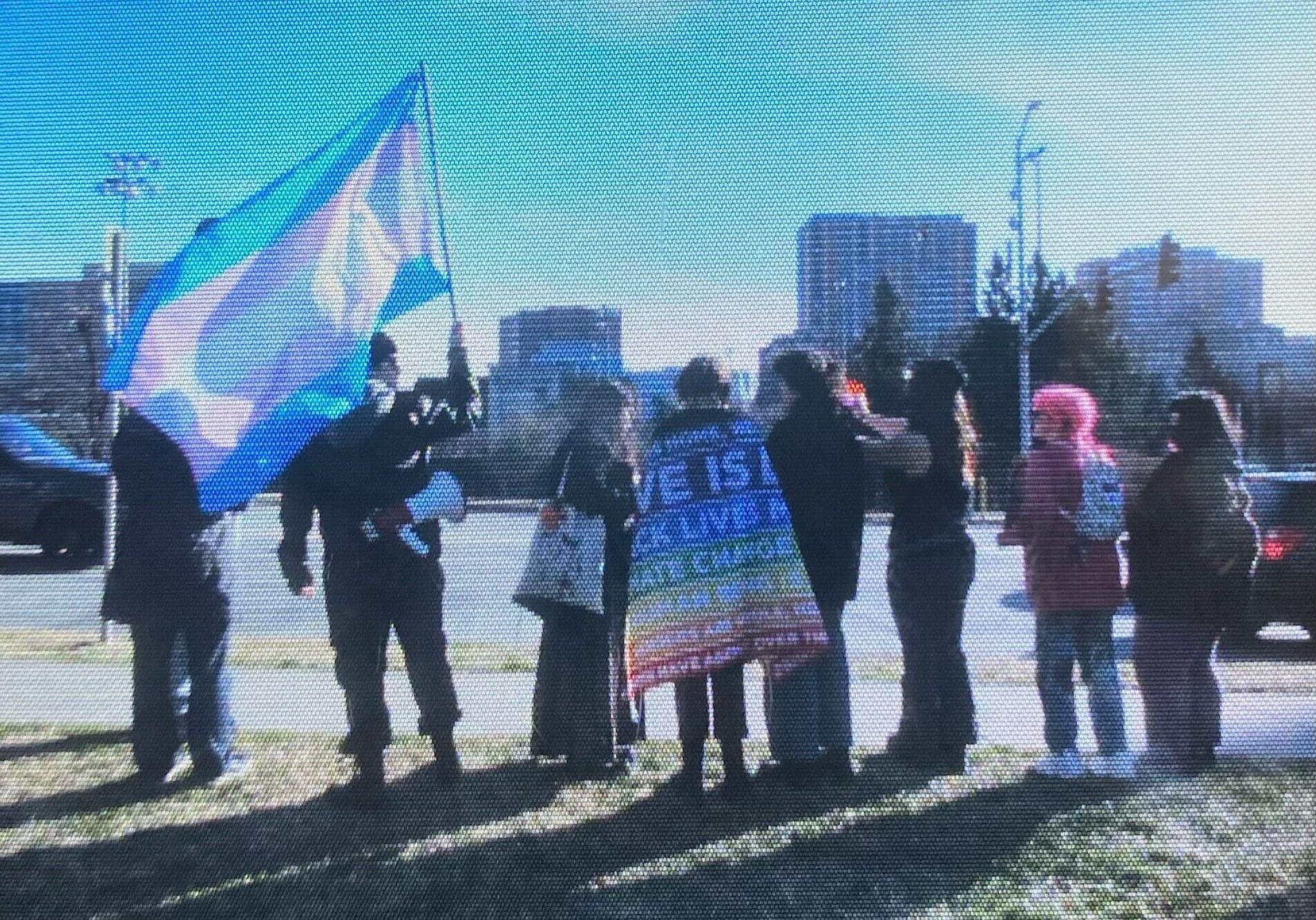 A group of high schoolers stand together, one holding a trans pride flag up in the air and a megaphone linked to their belt, another has a pride flat wrapped around their back with phrases such as "Love is Love" printed in white letters.