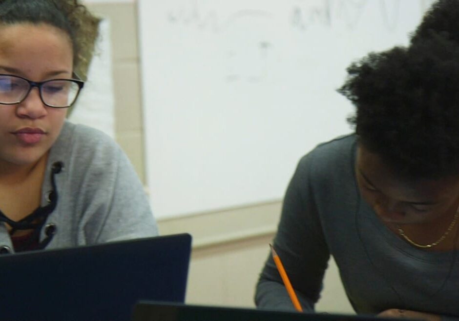 two young women at work, one on computer, one with a pencil