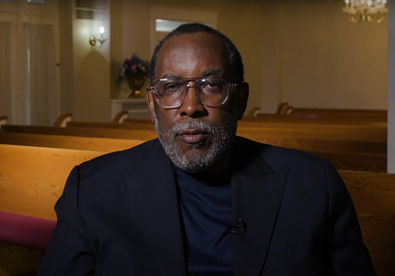 A man in a black suit with a beard and glasses sits in a pew in a chapel.