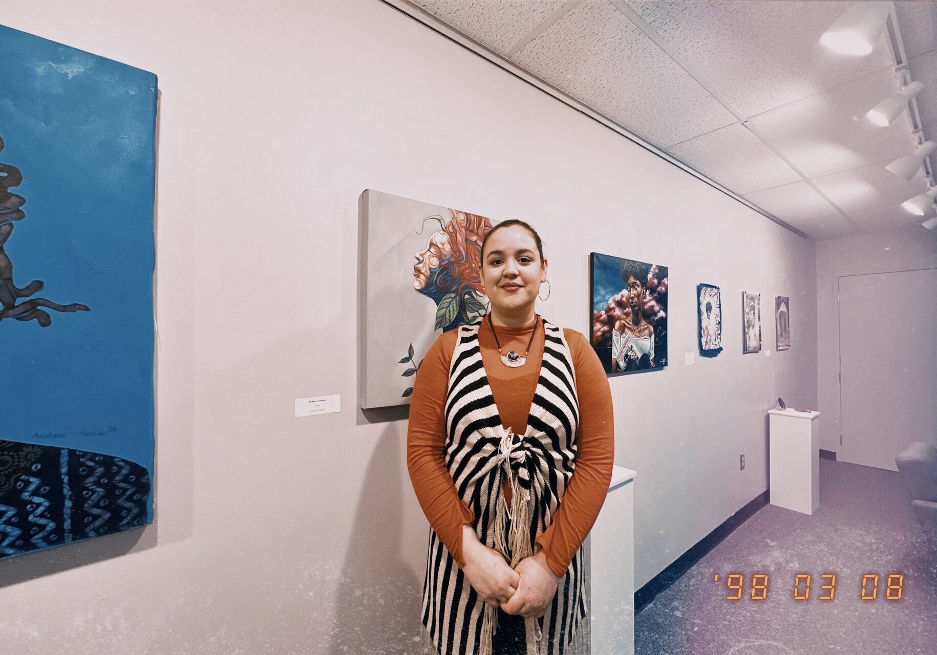 A woman wearing a striped shirt over an orange long sleeve shirt poses in an art gallery setting. Shai Perry is gallery coordinator at the Kansas City Kansas Community College. She is also an artist and is from Tennessee. Photo by Vicky Diaz-Camacho for Flatland.