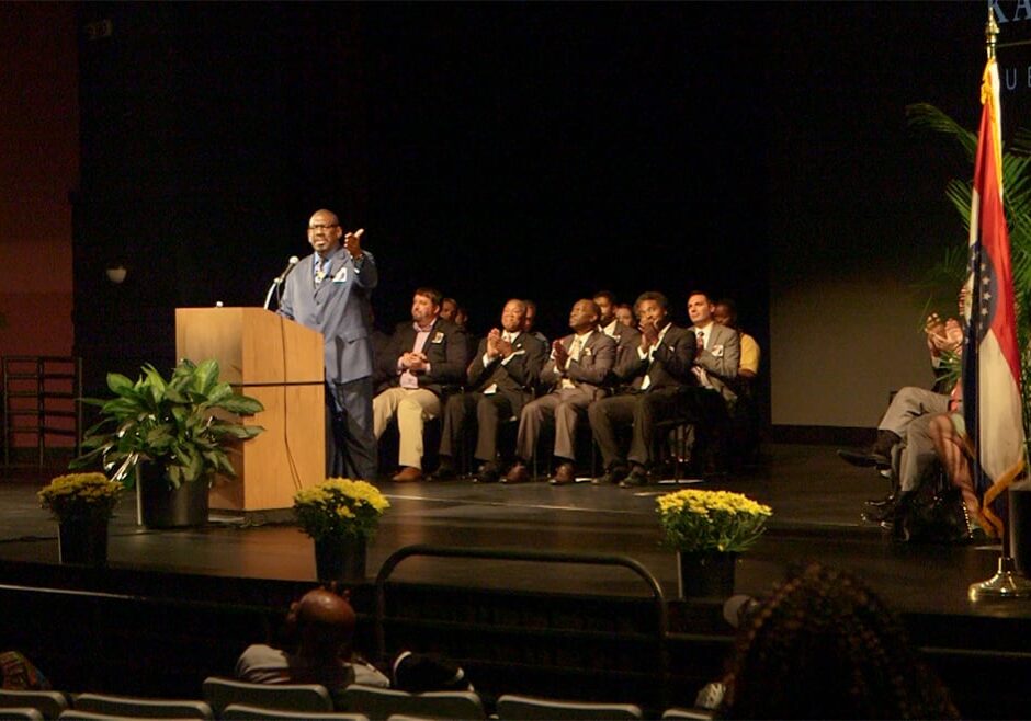 Man stands at podium on stage with a hand outstretched to the audience.