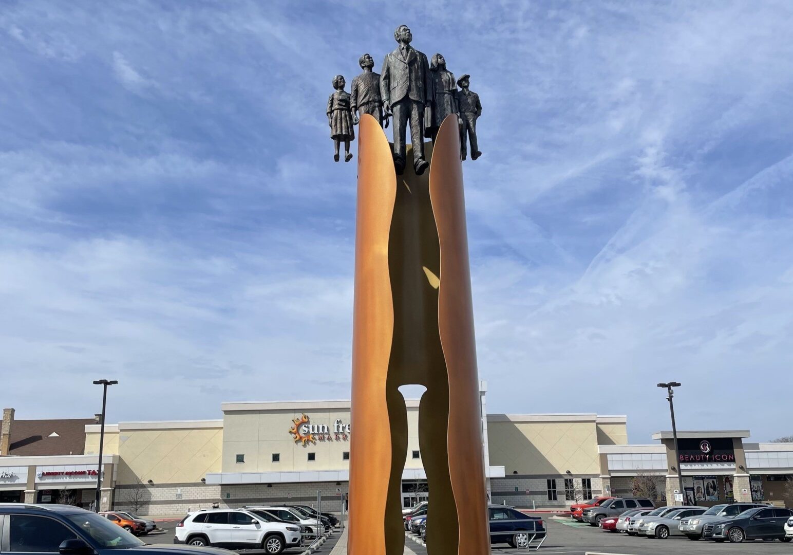 A tall bronze memorial with figures at the top in front of Sun Fresh market.