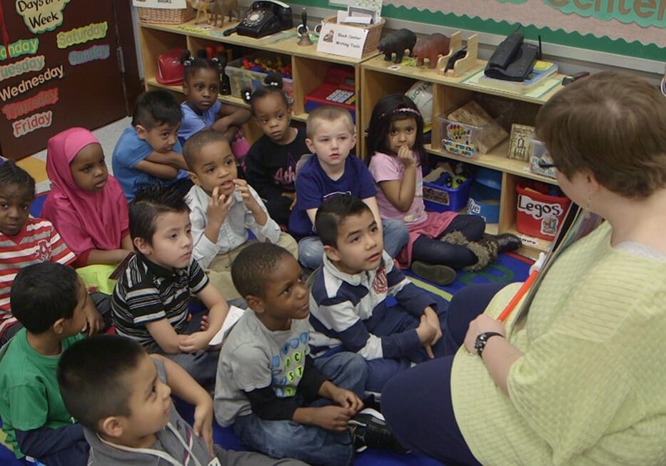 Picture of children sitting on the floor listening to teacher read book.