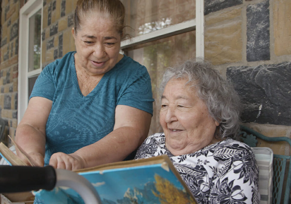 Woman sitting with older woman on porch, reading a book.