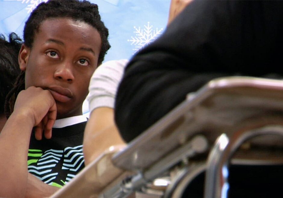Close-up of teenage, African-American male sitting in a desk and looking towards the front of the classroom, looking mildly bored.
