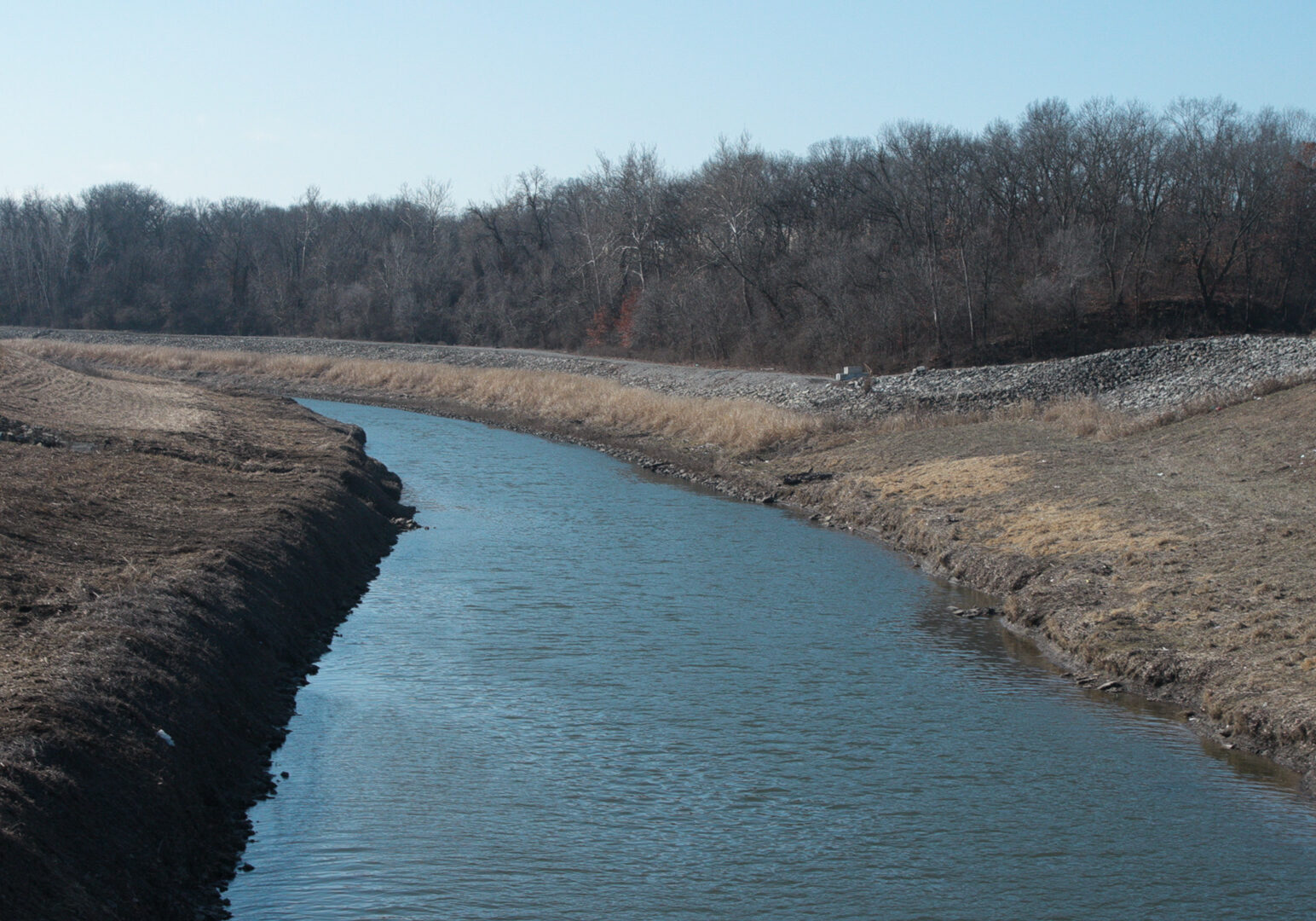 A photo of the Blue River. The river is larger at the foreground of the image and thins to a point in the background of the image. The water is blue with small ripples. On the sides of the river are clean, grassy banks. Just beyond the river bend in the background of the photo is a treeline.