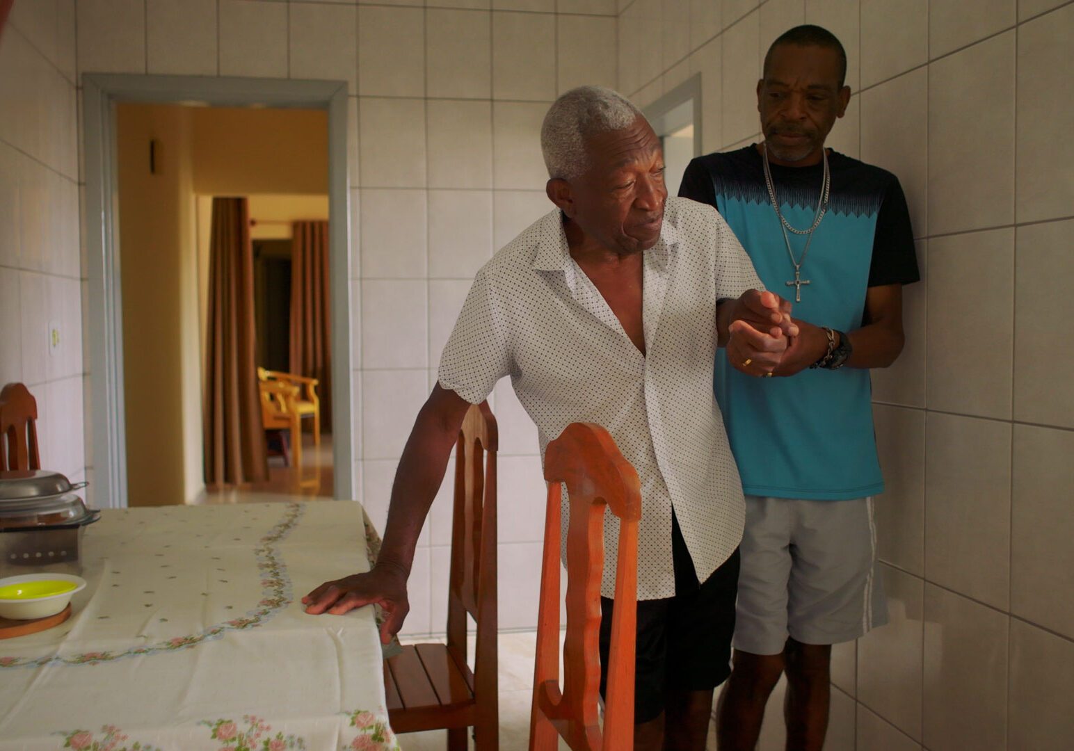 Younger man assists older man walking through dining room.