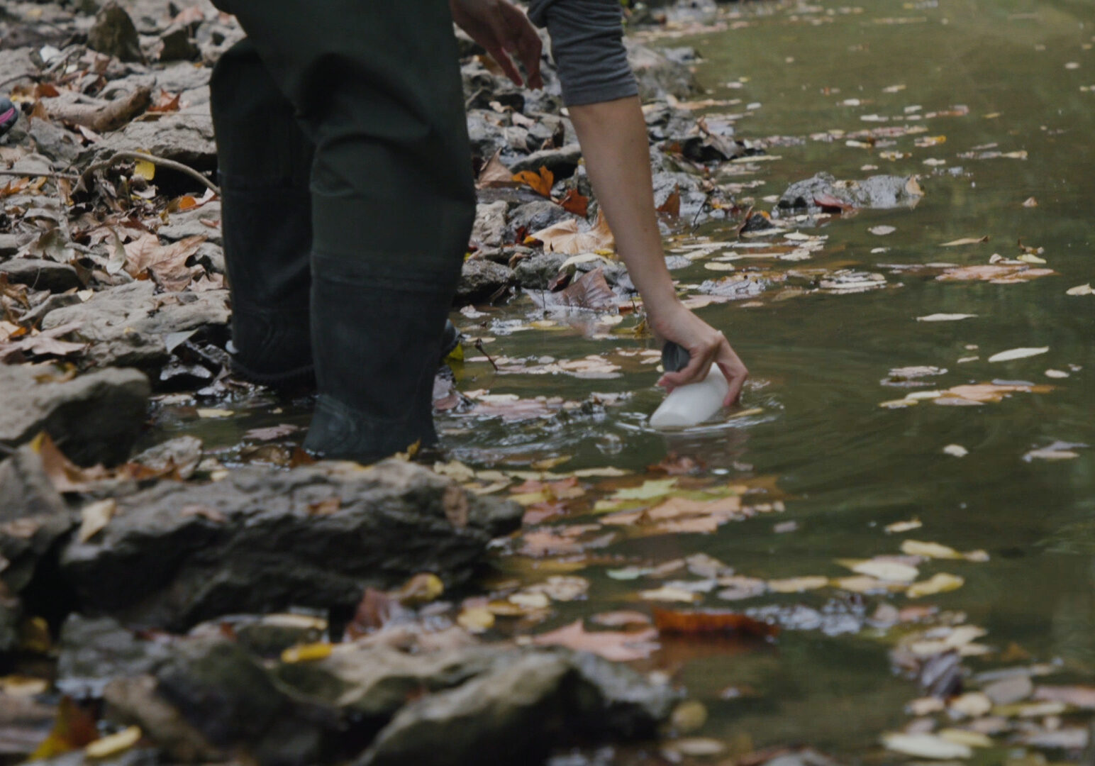 A person bends down and submurges a clear bottle into a green creek. They are wearing black boots. There are orange leaves scattered around them.