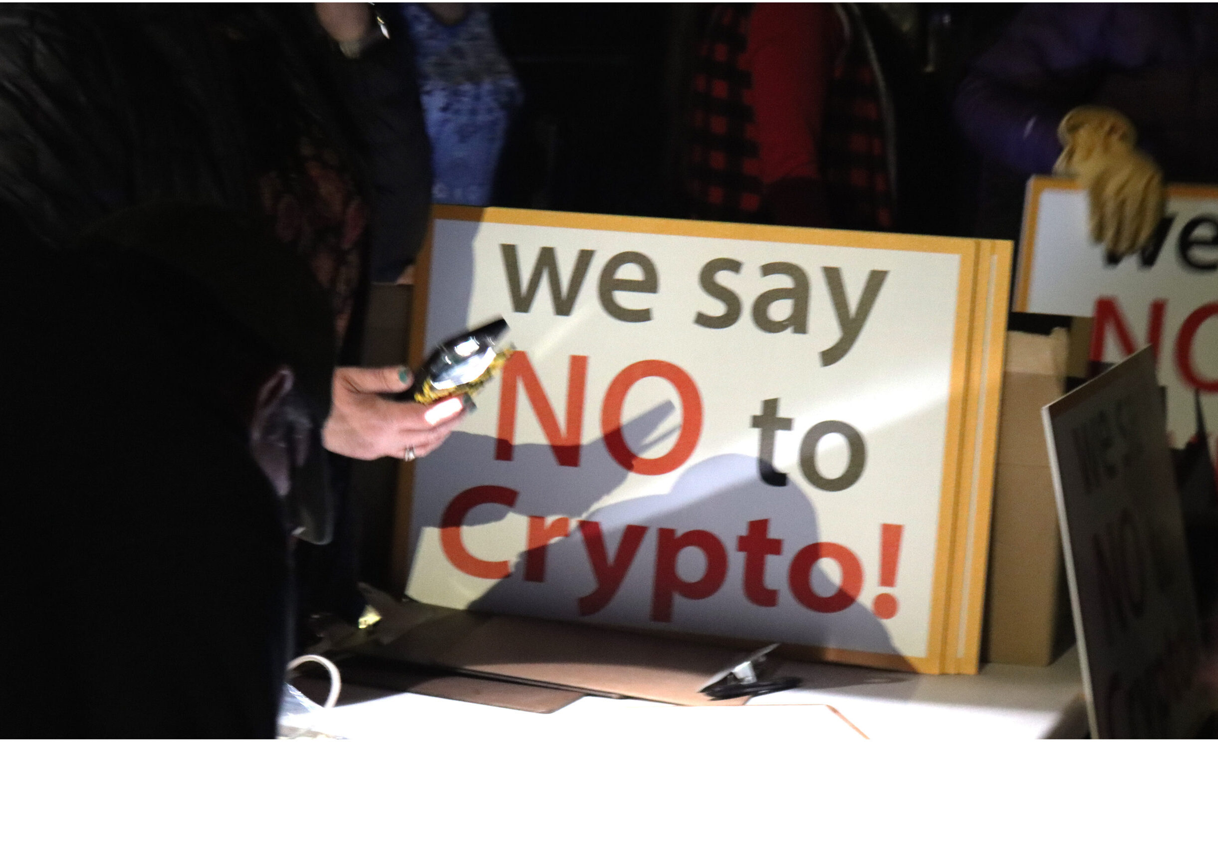 someone holds a phone with the flashlight on up to a table. Next to them sits a stack of signs that read "we say NO to Crypto"