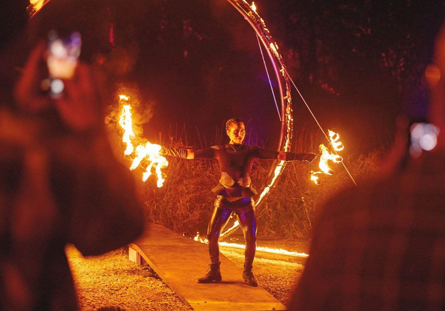 A fire performer in black stands waving flames in the dark forest.