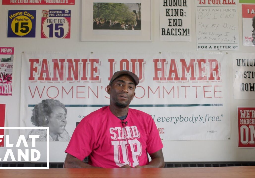 Man in red shirt that reads "stand up" sits at table with Fannie Lou Hamer board behind him.