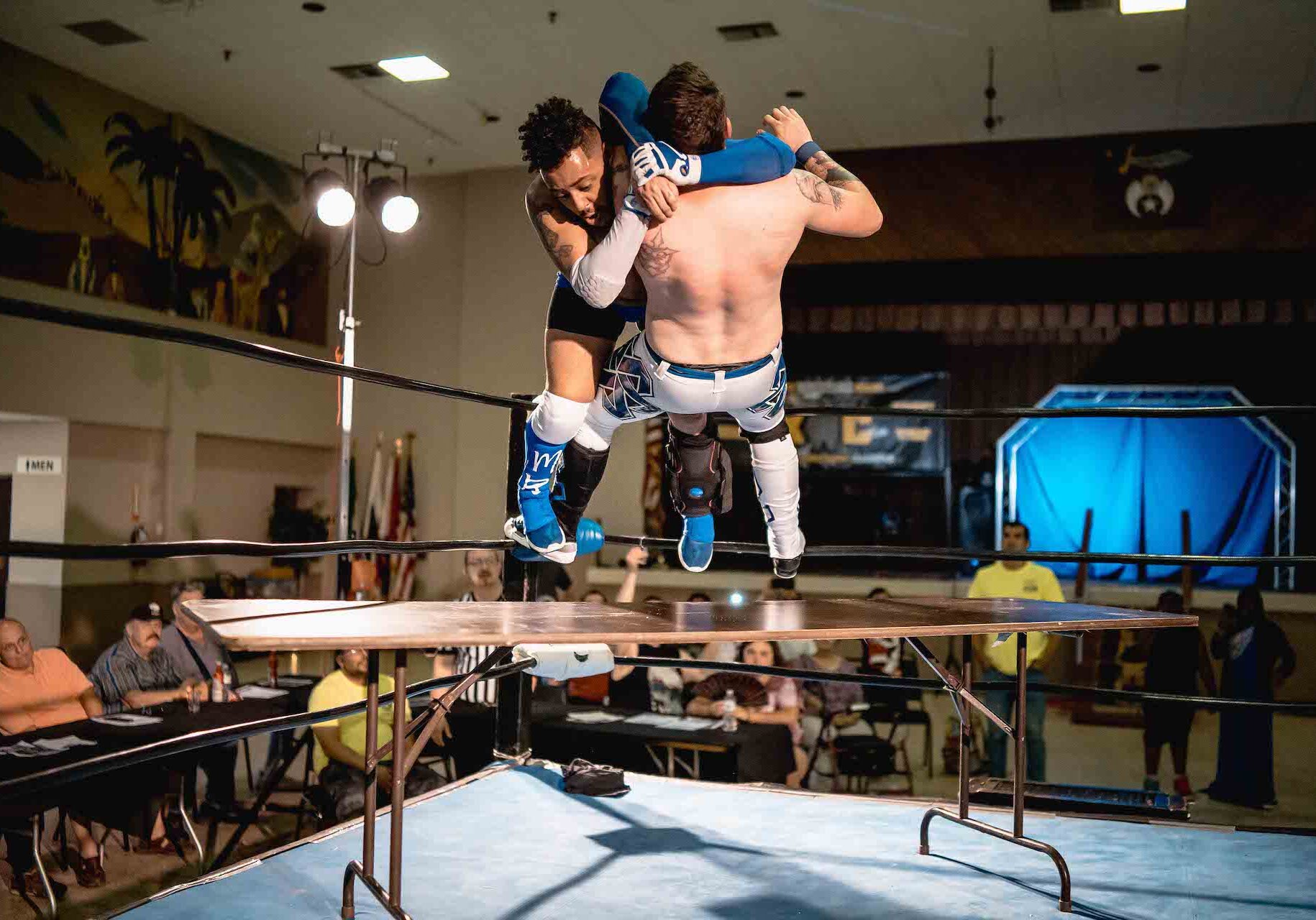A wrestler takes another wrestler off the turnbuckle and through a table.