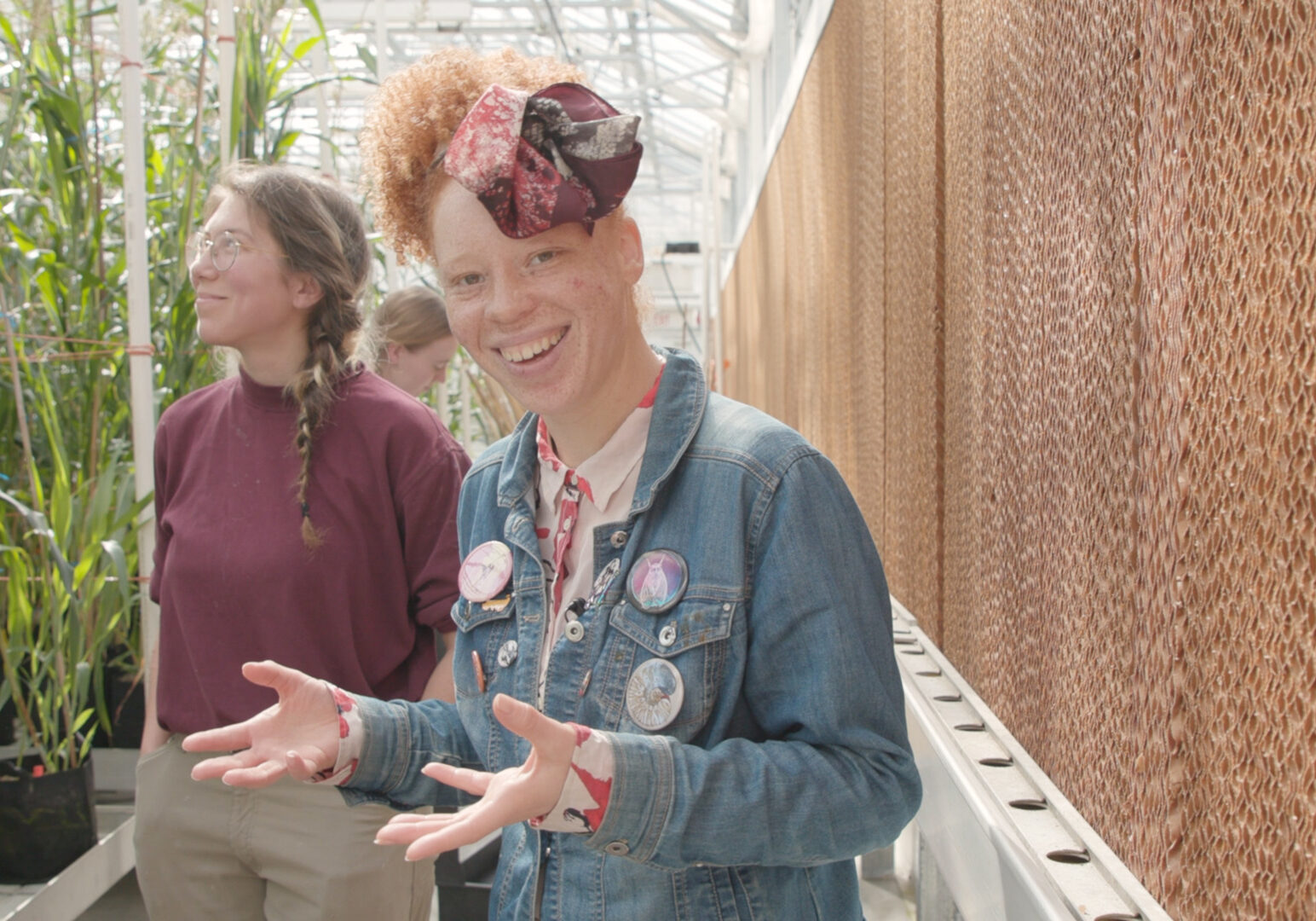 A woman with red hair and a scarf smiles and gestures with her hands. Behind her are rows of tall sorghum plants and a woman with a maroon tshirt.