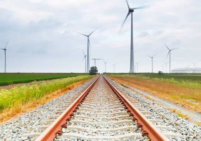 Train tracks against a backdrop of green grass and wind turbines illustrates a new project called the Grain Belt Express. This new effort is a "massive high-powered transmission line delivering renewable energy."