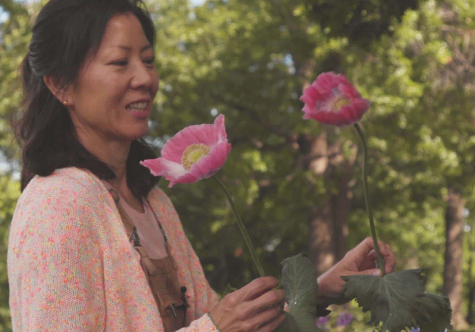 A woman holds two tall-stemmed pink flowers and smiles.