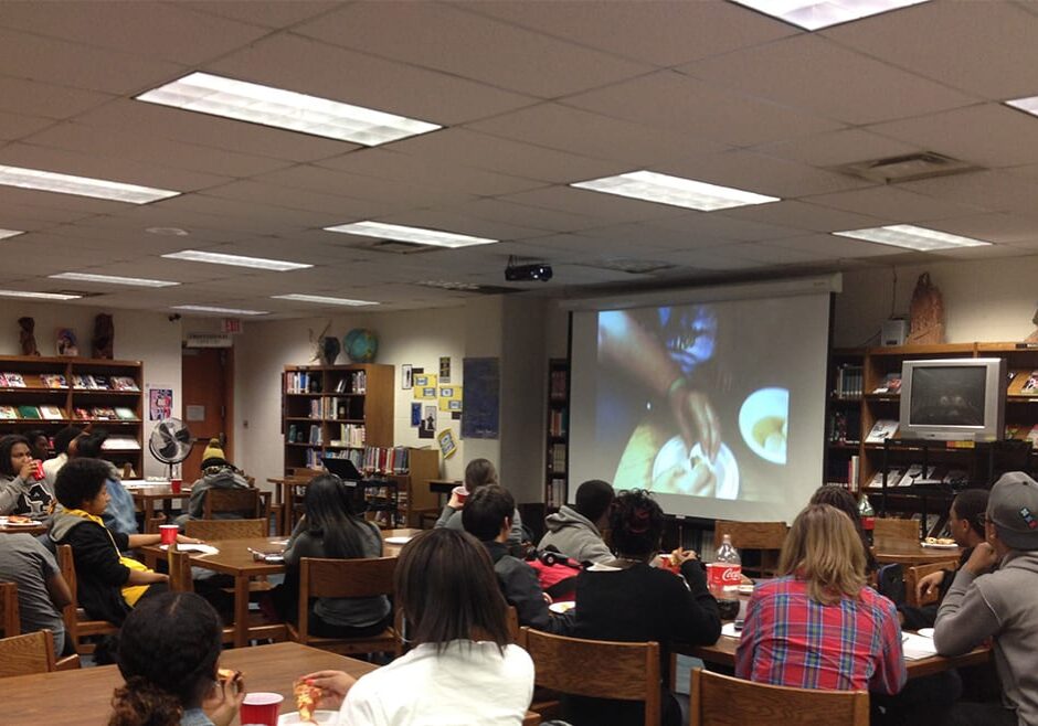 Group of high school students sit in library and watch film on large project screen.