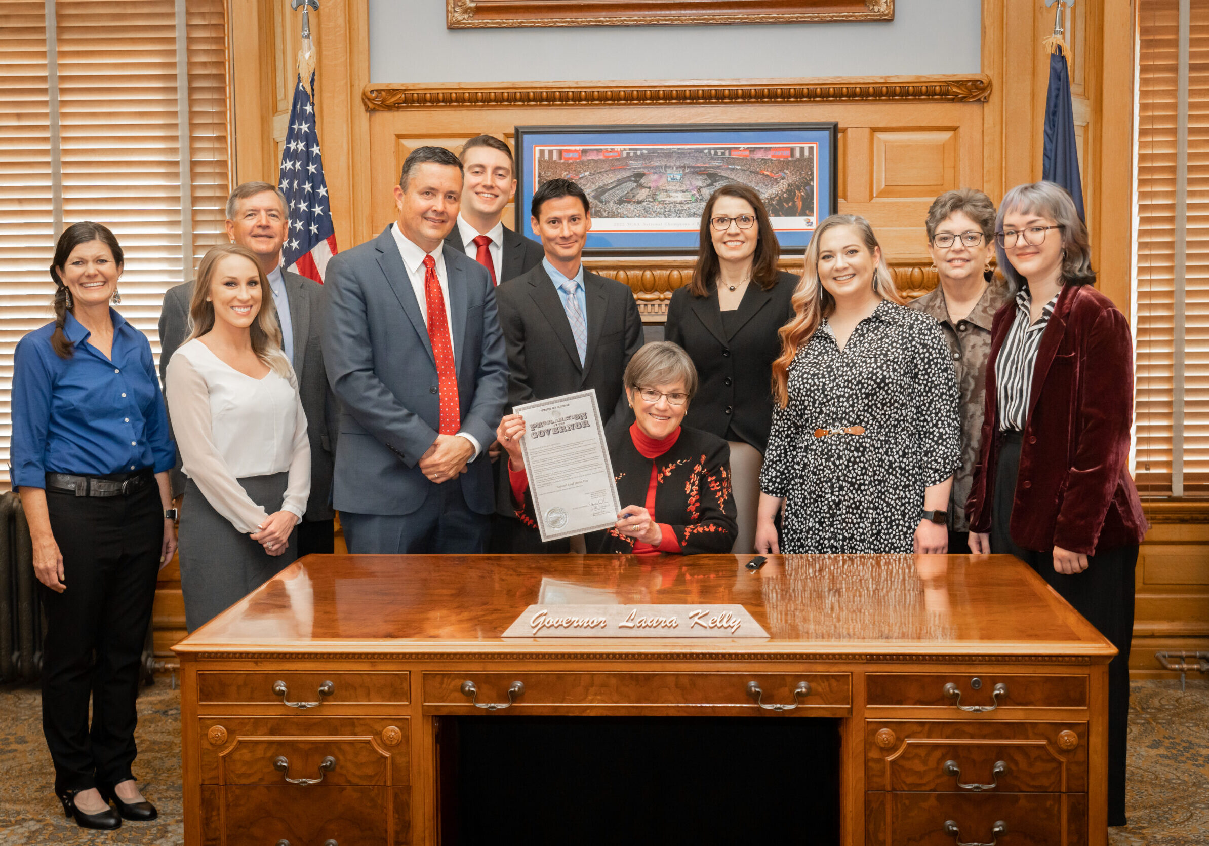 Group of people stand around KS Governor Laura Kelly at her desk. Kelly holds a document proclaiming rural national health day in Kansas.