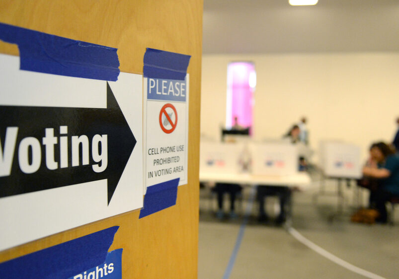 A wooden door opens into a room with blurred figures, white walls and voter dividers sitting on tables. On the door is a sign that has a black arrow and the word Voting. It also has a sign with a red circle with hash through it around a cell phone that says, "cell phone use is prohibited".