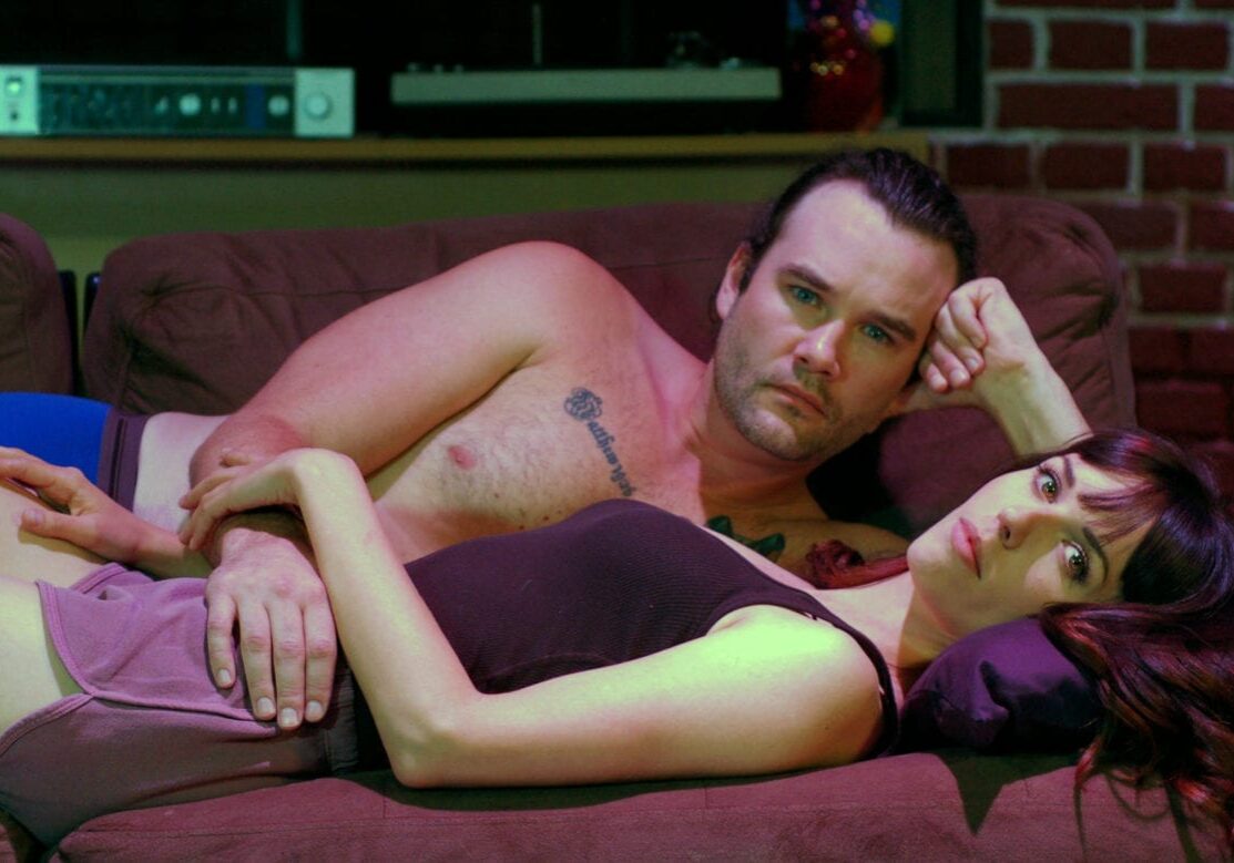 A man and woman laying on a sofa as part of a play, "The Way We Get By."