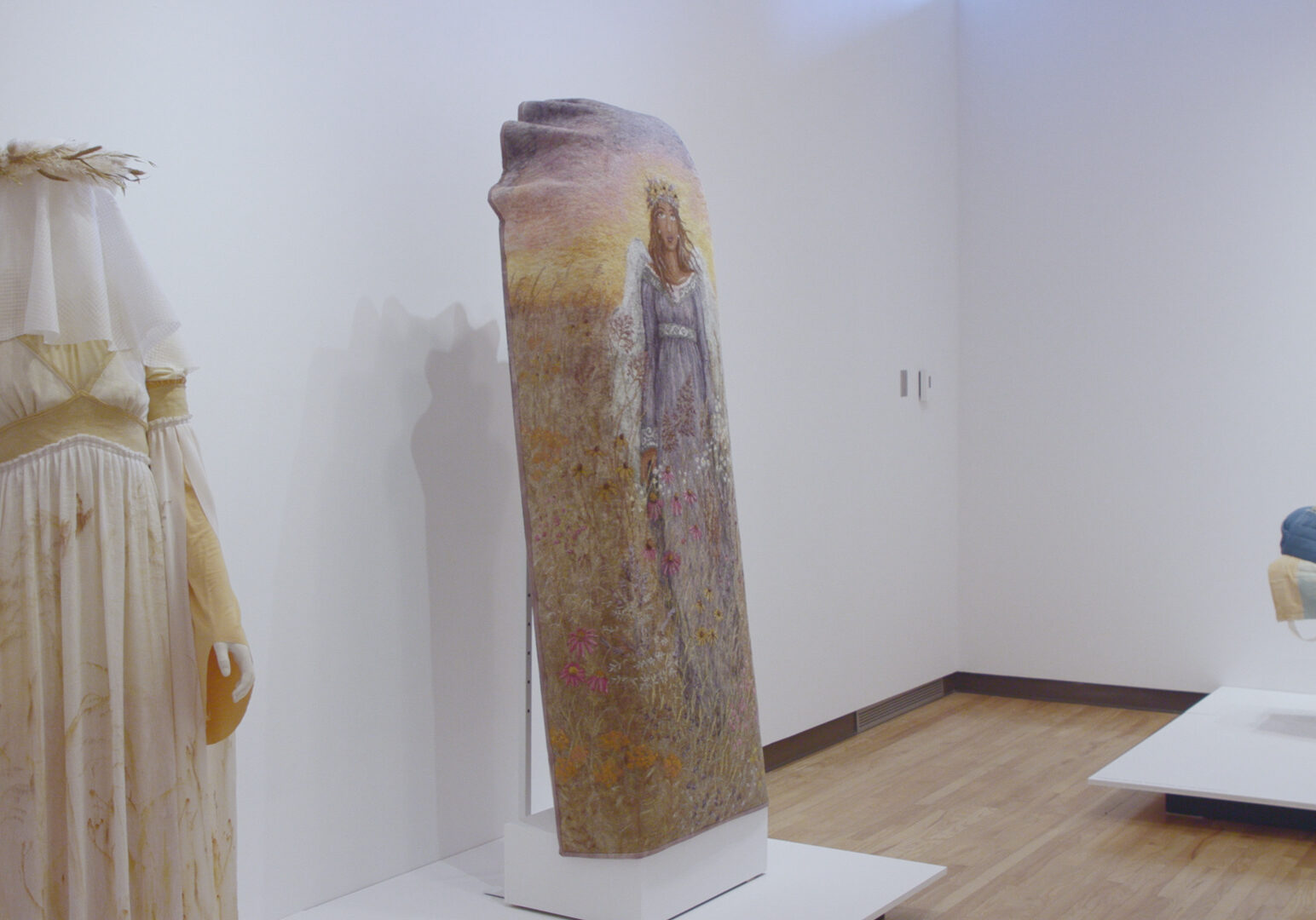 A photo of a museum exhibition. At the left of the photo, a mannequin stands with a light yellow gown and a white veil over its head. The veil is peppered with light brown floral prints. On top of the mannequins head is a crown of prairie grass. To the right of the mannequin with the gown, and in the center of the photo is a coffin cover. The cover depicts an angel in a blue gown with white wings. She is depicted standing in the prairie, covered in prairie flowers. Off to the right side of the photograph is a mannequin lying flat, covered in a blue burial garment.