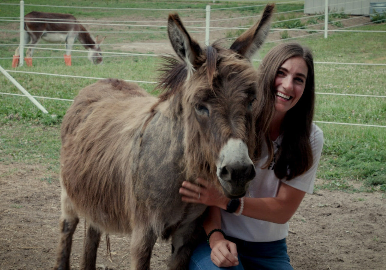 A woman with dark hair crouches next to a short long haired donkey. She laughs while petting his beard.