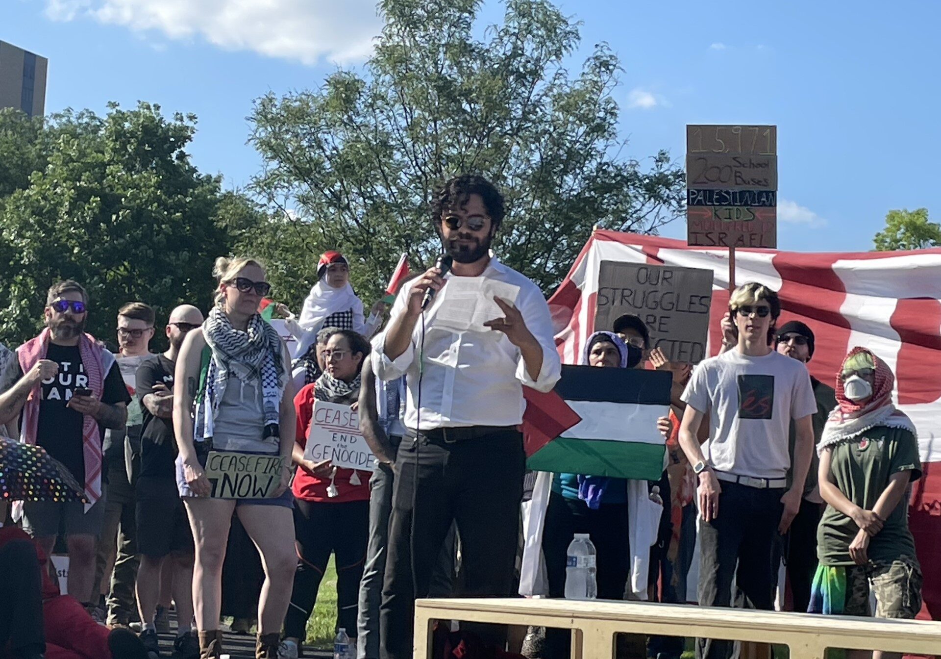 people standing outdoors. banners, palestinian flag, man reading from a white paper