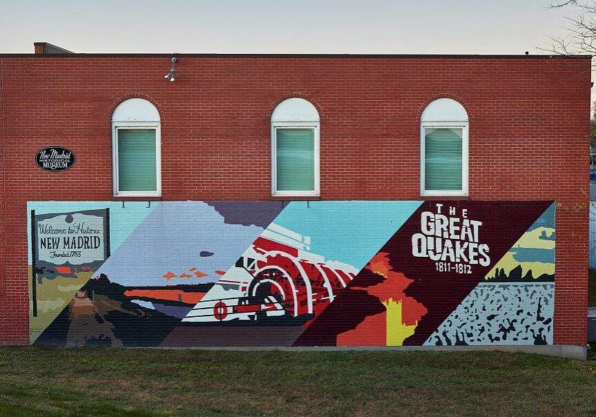 A colorful mural on the side of a brick building with the words "Welcome to Historic New Madrid" and "The Great Quakes" at dusk