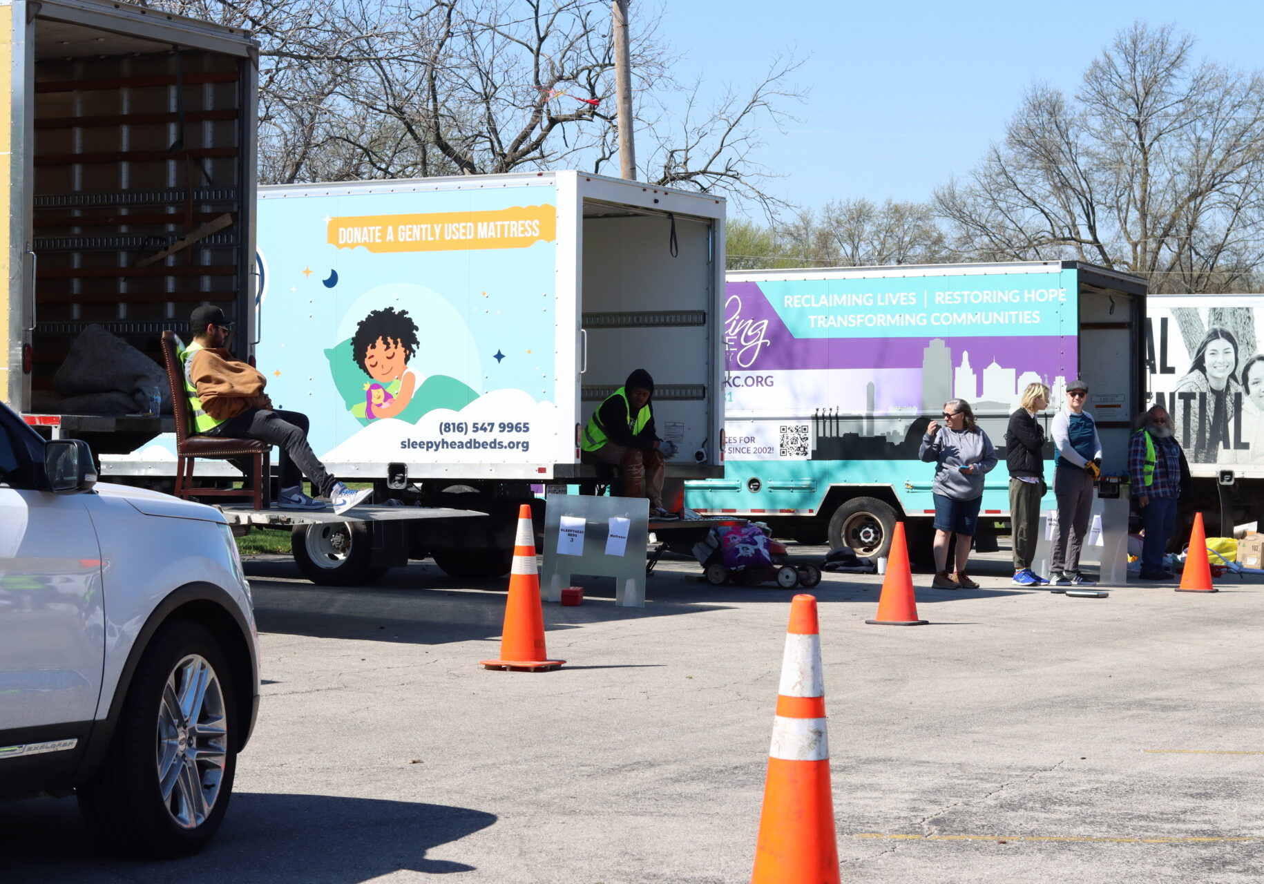 A row of box trucks with various nonprofit logos are lined up in a parking lot. A car drives down a row of cones behind the trucks while volunteers stand nearby in yellow vests.