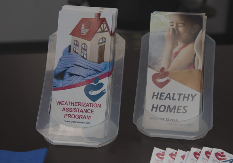 Two stacks of pamphlets on a table. The table is brown. The pamphlets to the left of the screen advertise a weatherization assistance program. These pamphlets have a house with a red roof on them wrapped in a blue scarf. The text is bold and red. The pamphlets on the right advertise the healthy homes program. On these pamphlets, a young girl holds her nose. The text on these pamphlets is black and bold.